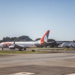 Aviões comerciais taxiando em um aeroporto sob céu ensolarado, antes da decolagem.