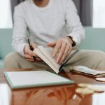 Pessoa analisa as próprias finanças em uma mesa, com caderno, dinheiro e laptop.