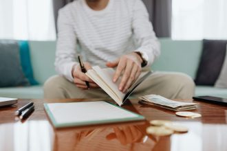 Pessoa analisa as próprias finanças em uma mesa, com caderno, dinheiro e laptop.