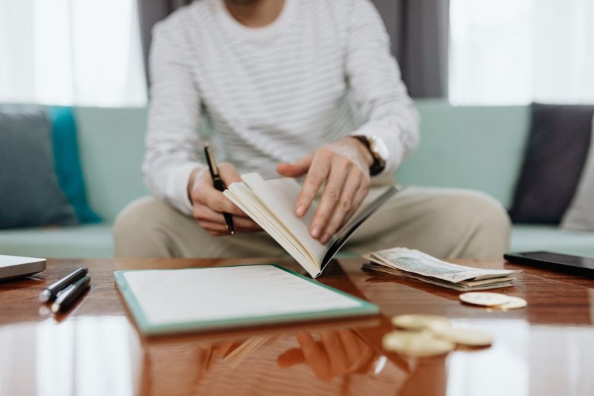 Pessoa analisa as próprias finanças em uma mesa, com caderno, dinheiro e laptop.