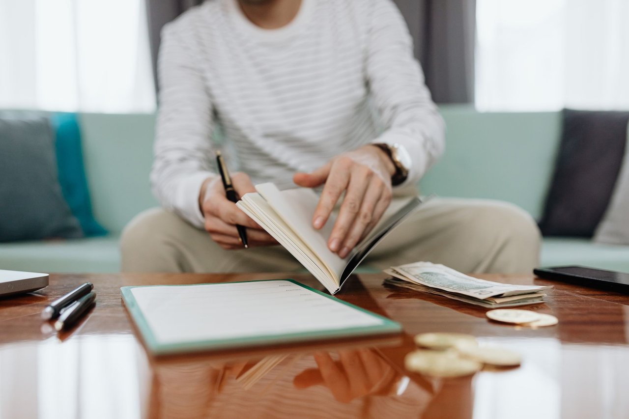 Pessoa analisa as próprias finanças em uma mesa, com caderno, dinheiro e laptop.